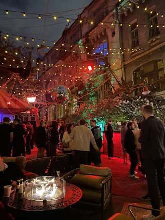 An outdoor social gathering taking place at night, set against a backdrop of a multi-story building with decorative lighting. The area is adorned with string lights and a disco ball, creating a festive atmosphere. A group of people are mingling and chatting, with some seated around a table with a circular fire pit. The lighting gives the scene a warm glow with reds and greens, highlighting the vibrant atmosphere.