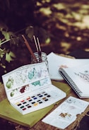 A set of watercolor paints is open on top of a green book, with various colors splattered around the palette. Paintbrushes are standing inside a glass jar, and a sketchbook with floral drawings is open nearby. The setting appears to be outdoors, with blurred greenery in the background.