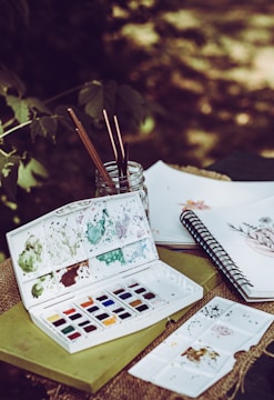 A set of watercolor paints is open on top of a green book, with various colors splattered around the palette. Paintbrushes are standing inside a glass jar, and a sketchbook with floral drawings is open nearby. The setting appears to be outdoors, with blurred greenery in the background.