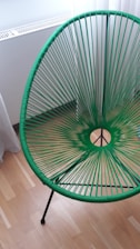 A modern chair with a circular woven design in vibrant green is displayed on a wooden floor near a white wall and radiator. The chair features a tripod-style base.