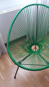 A modern chair with a circular woven design in vibrant green is displayed on a wooden floor near a white wall and radiator. The chair features a tripod-style base.