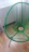 A modern chair with a circular woven design in vibrant green is displayed on a wooden floor near a white wall and radiator. The chair features a tripod-style base.