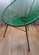 A green, modern woven chair with a circular pattern in its seating area. It has sleek, black metal legs and is set against a light wooden floor with a white curtain in the background.