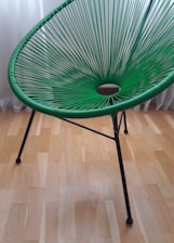 A green, modern woven chair with a circular pattern in its seating area. It has sleek, black metal legs and is set against a light wooden floor with a white curtain in the background.