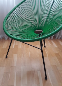 A green, modern woven chair with a circular pattern in its seating area. It has sleek, black metal legs and is set against a light wooden floor with a white curtain in the background.