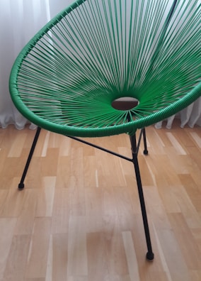 A green, modern woven chair with a circular pattern in its seating area. It has sleek, black metal legs and is set against a light wooden floor with a white curtain in the background.
