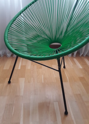 A green, modern woven chair with a circular pattern in its seating area. It has sleek, black metal legs and is set against a light wooden floor with a white curtain in the background.