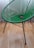 A green, modern woven chair with a circular pattern in its seating area. It has sleek, black metal legs and is set against a light wooden floor with a white curtain in the background.