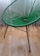 A green, modern woven chair with a circular pattern in its seating area. It has sleek, black metal legs and is set against a light wooden floor with a white curtain in the background.