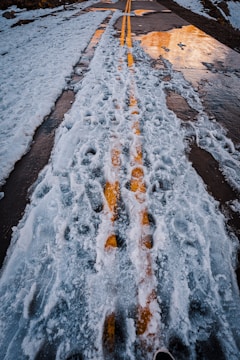 A road covered in snow and ice with visible yellow lines running through the center. The sunlight reflects off the wet surface, creating a mixture of blue, white, and yellow hues. The surrounding area shows patches of snow and some exposed asphalt.