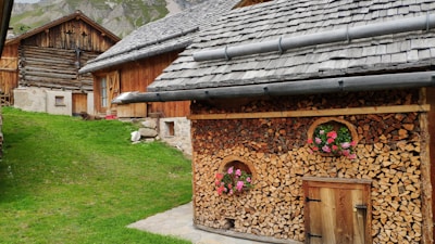 Charming rustic wooden houses nestled in a mountainous setting, featuring neatly stacked firewood walls adorned with pink flowers in small circular window-like openings. A green grass lawn creates a natural contrast, while the backdrop reveals rugged mountain peaks.