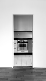 A monochrome image of a kitchen featuring a stainless steel oven. The kitchen is observed through an open doorway with a view of tiled flooring extending to a hardwood floor in the foreground. Sleek, modern cabinets with horizontal handles are situated above and beside the oven.