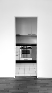 A monochrome image of a kitchen featuring a stainless steel oven. The kitchen is observed through an open doorway with a view of tiled flooring extending to a hardwood floor in the foreground. Sleek, modern cabinets with horizontal handles are situated above and beside the oven.