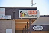 A sign for a coin-operated laundry is prominently displayed, featuring an illustration of a person doing laundry. Next to it is a sign for a tattoo shop named Rollin Gypsy Tattoo. Various payment methods accepted are shown on another sign, including EBT and major credit cards. An overhang and a satellite dish are visible on the building's roof.