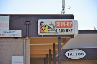 A sign for a coin-operated laundry is prominently displayed, featuring an illustration of a person doing laundry. Next to it is a sign for a tattoo shop named Rollin Gypsy Tattoo. Various payment methods accepted are shown on another sign, including EBT and major credit cards. An overhang and a satellite dish are visible on the building's roof.