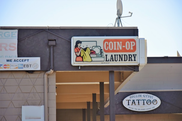 A sign for a coin-operated laundry is prominently displayed, featuring an illustration of a person doing laundry. Next to it is a sign for a tattoo shop named Rollin Gypsy Tattoo. Various payment methods accepted are shown on another sign, including EBT and major credit cards. An overhang and a satellite dish are visible on the building's roof.