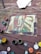 An abstract painting on an old wooden surface, possibly a deck. The painting features a face with various colors, including red, blue, and green brushstrokes. Surrounding the canvas are several open paint cans with brushes, displaying colors such as red, green, yellow, and white. A rolled-up piece of fabric or paper lies to the side.