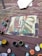An abstract painting on an old wooden surface, possibly a deck. The painting features a face with various colors, including red, blue, and green brushstrokes. Surrounding the canvas are several open paint cans with brushes, displaying colors such as red, green, yellow, and white. A rolled-up piece of fabric or paper lies to the side.