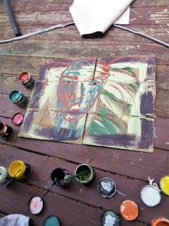 An abstract painting on an old wooden surface, possibly a deck. The painting features a face with various colors, including red, blue, and green brushstrokes. Surrounding the canvas are several open paint cans with brushes, displaying colors such as red, green, yellow, and white. A rolled-up piece of fabric or paper lies to the side.