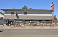 A two-story commercial building with a gray exterior, located beside a street. The storefront is protected with a chain-link fence, behind which various tools and equipment like ladders, bicycles, and lawnmowers are displayed. A person is standing near the entrance. Red signage on the right corner indicates 'Loans' and 'Top Kick.'