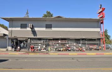 A two-story commercial building with a gray exterior, located beside a street. The storefront is protected with a chain-link fence, behind which various tools and equipment like ladders, bicycles, and lawnmowers are displayed. A person is standing near the entrance. Red signage on the right corner indicates 'Loans' and 'Top Kick.'