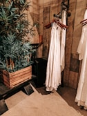 An interior setting features wooden walls and flooring, with a leafy green plant in a rustic wooden planter. Nearby, a series of white garments are hung on wooden hangers attached to a metal pipe rack. The setting has a warm, earthy aesthetic.