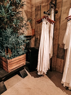 An interior setting features wooden walls and flooring, with a leafy green plant in a rustic wooden planter. Nearby, a series of white garments are hung on wooden hangers attached to a metal pipe rack. The setting has a warm, earthy aesthetic.