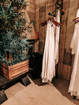 An interior setting features wooden walls and flooring, with a leafy green plant in a rustic wooden planter. Nearby, a series of white garments are hung on wooden hangers attached to a metal pipe rack. The setting has a warm, earthy aesthetic.