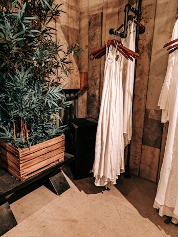 An interior setting features wooden walls and flooring, with a leafy green plant in a rustic wooden planter. Nearby, a series of white garments are hung on wooden hangers attached to a metal pipe rack. The setting has a warm, earthy aesthetic.