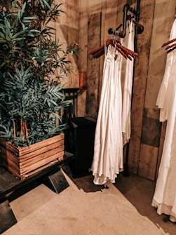 An interior setting features wooden walls and flooring, with a leafy green plant in a rustic wooden planter. Nearby, a series of white garments are hung on wooden hangers attached to a metal pipe rack. The setting has a warm, earthy aesthetic.