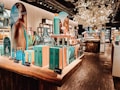 A well-lit interior of a cosmetics store featuring a wooden display table with vibrant blue and turquoise boxed skincare products. There are promotional signs showcasing skincare items, decorative large glass jars, and an arrangement of white artificial blossoms, creating a luxurious atmosphere. Shelves in the background are filled with various beauty products.