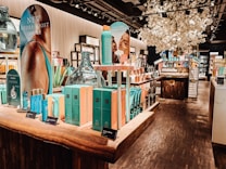 A well-lit interior of a cosmetics store featuring a wooden display table with vibrant blue and turquoise boxed skincare products. There are promotional signs showcasing skincare items, decorative large glass jars, and an arrangement of white artificial blossoms, creating a luxurious atmosphere. Shelves in the background are filled with various beauty products.
