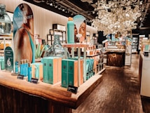 A well-lit interior of a cosmetics store featuring a wooden display table with vibrant blue and turquoise boxed skincare products. There are promotional signs showcasing skincare items, decorative large glass jars, and an arrangement of white artificial blossoms, creating a luxurious atmosphere. Shelves in the background are filled with various beauty products.