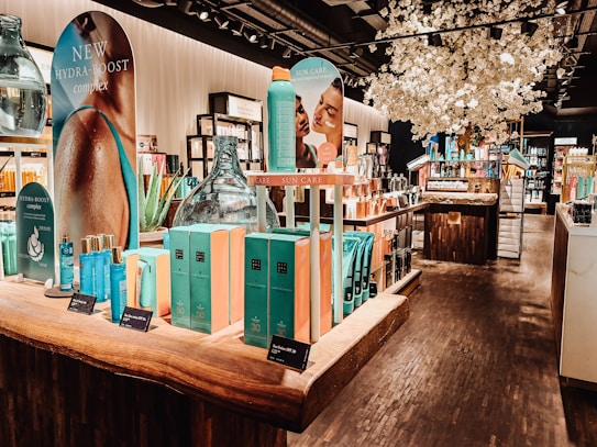 A well-lit interior of a cosmetics store featuring a wooden display table with vibrant blue and turquoise boxed skincare products. There are promotional signs showcasing skincare items, decorative large glass jars, and an arrangement of white artificial blossoms, creating a luxurious atmosphere. Shelves in the background are filled with various beauty products.