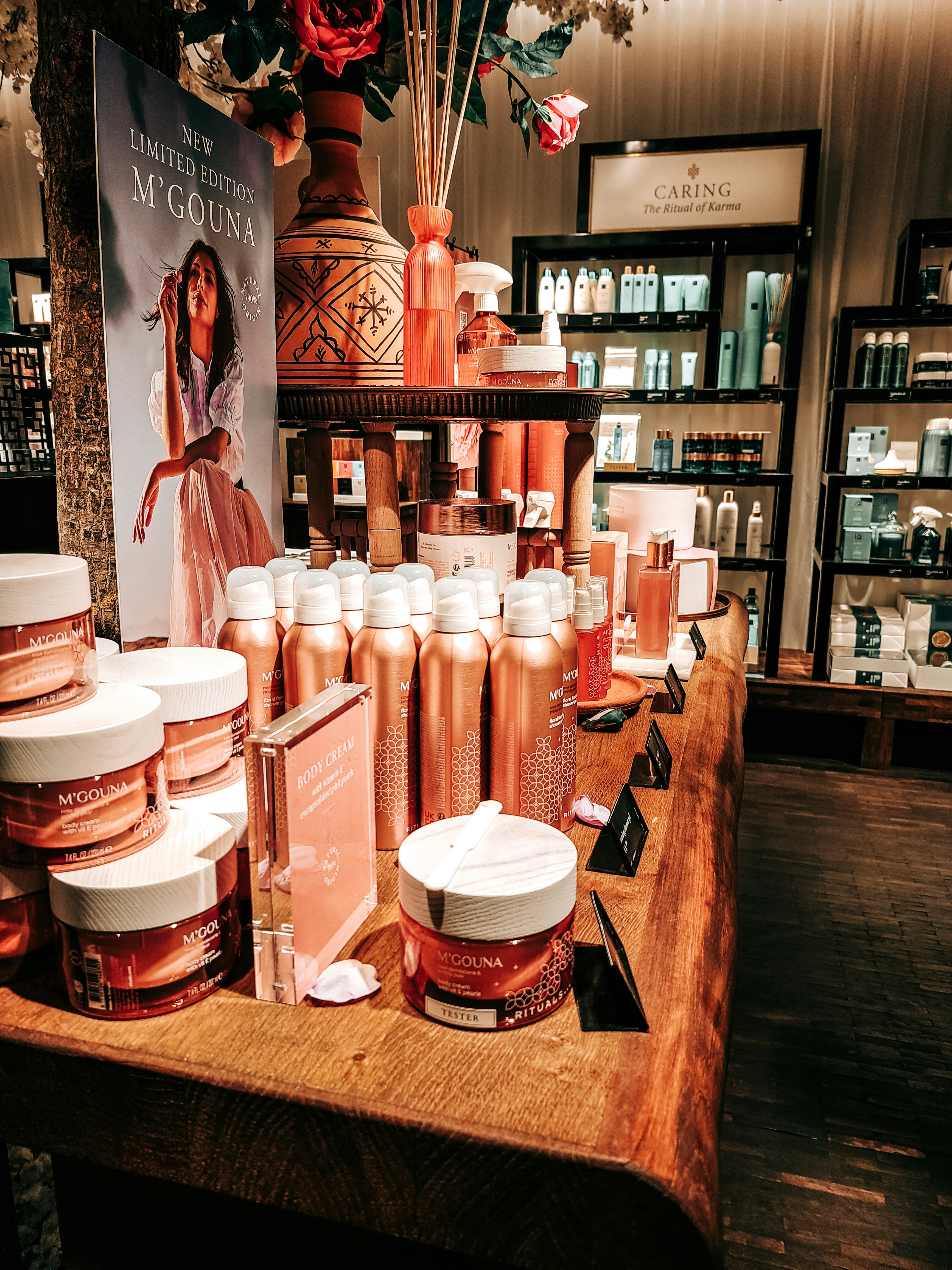 An elegant display in a store features an array of beauty products, including body creams and lotions, set on a wooden table. The product packaging is in shades of pink and peach. A decorative vase with bamboo sticks and rose petals is placed among the products. Advertisements for a new limited edition line are visible, with a poster featuring a woman in a flowing dress. Shelves filled with various skincare products line the walls in the background.