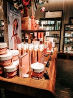 An elegant display in a store features an array of beauty products, including body creams and lotions, set on a wooden table. The product packaging is in shades of pink and peach. A decorative vase with bamboo sticks and rose petals is placed among the products. Advertisements for a new limited edition line are visible, with a poster featuring a woman in a flowing dress. Shelves filled with various skincare products line the walls in the background.