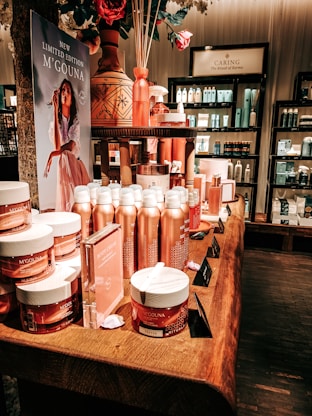 An elegant display in a store features an array of beauty products, including body creams and lotions, set on a wooden table. The product packaging is in shades of pink and peach. A decorative vase with bamboo sticks and rose petals is placed among the products. Advertisements for a new limited edition line are visible, with a poster featuring a woman in a flowing dress. Shelves filled with various skincare products line the walls in the background.