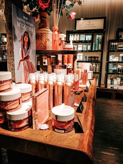 An elegant display in a store features an array of beauty products, including body creams and lotions, set on a wooden table. The product packaging is in shades of pink and peach. A decorative vase with bamboo sticks and rose petals is placed among the products. Advertisements for a new limited edition line are visible, with a poster featuring a woman in a flowing dress. Shelves filled with various skincare products line the walls in the background.