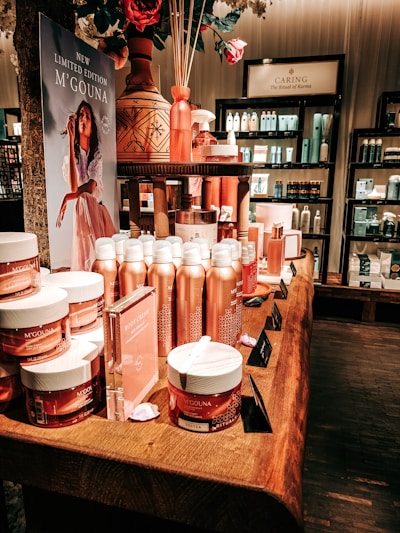 An elegant display in a store features an array of beauty products, including body creams and lotions, set on a wooden table. The product packaging is in shades of pink and peach. A decorative vase with bamboo sticks and rose petals is placed among the products. Advertisements for a new limited edition line are visible, with a poster featuring a woman in a flowing dress. Shelves filled with various skincare products line the walls in the background.