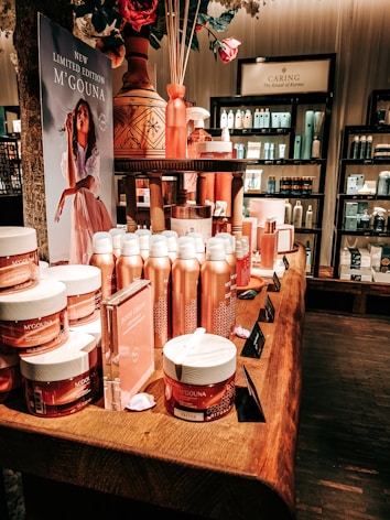 An elegant display in a store features an array of beauty products, including body creams and lotions, set on a wooden table. The product packaging is in shades of pink and peach. A decorative vase with bamboo sticks and rose petals is placed among the products. Advertisements for a new limited edition line are visible, with a poster featuring a woman in a flowing dress. Shelves filled with various skincare products line the walls in the background.