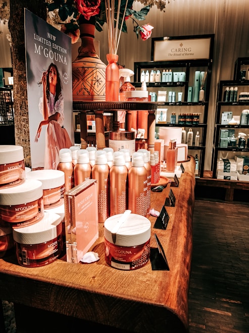 An elegant display in a store features an array of beauty products, including body creams and lotions, set on a wooden table. The product packaging is in shades of pink and peach. A decorative vase with bamboo sticks and rose petals is placed among the products. Advertisements for a new limited edition line are visible, with a poster featuring a woman in a flowing dress. Shelves filled with various skincare products line the walls in the background.