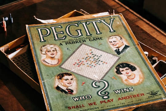 A vintage board game box with the title 'Pegity' prominently displayed. It features illustrations of four people, two men and two women, set in a retro style. The board inside the box is visible along with colored pegs. The phrase 'Who Wins' and 'Shall We Play Another' are included on the cover.