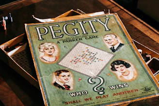 A vintage board game box with the title 'Pegity' prominently displayed. It features illustrations of four people, two men and two women, set in a retro style. The board inside the box is visible along with colored pegs. The phrase 'Who Wins' and 'Shall We Play Another' are included on the cover.