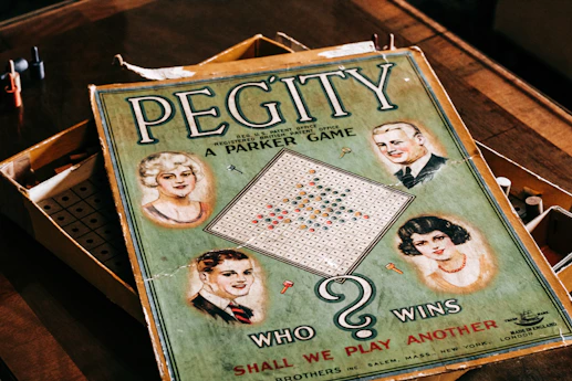 A vintage board game box with the title 'Pegity' prominently displayed. It features illustrations of four people, two men and two women, set in a retro style. The board inside the box is visible along with colored pegs. The phrase 'Who Wins' and 'Shall We Play Another' are included on the cover.