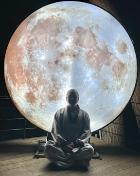 A person dressed in traditional attire sits in a meditative pose on a wooden floor, silhouetted against a large, illuminated image of the moon. The setting appears serene and contemplative, with the moon's detailed surface providing atmospheric lighting.