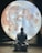 A person dressed in traditional attire sits in a meditative pose on a wooden floor, silhouetted against a large, illuminated image of the moon. The setting appears serene and contemplative, with the moon's detailed surface providing atmospheric lighting.