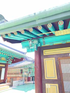 A traditional building with intricate wooden decorations and colorful patterns, featuring geometric and floral designs. The roof is adorned with multiple hues of green and blue, while the walls have accents of red and yellow. Detailed lattice work is visible on the doors, and the architectural style is reminiscent of Korean craftsmanship.