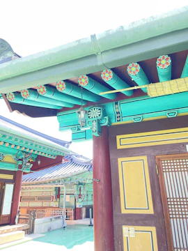 A traditional building with intricate wooden decorations and colorful patterns, featuring geometric and floral designs. The roof is adorned with multiple hues of green and blue, while the walls have accents of red and yellow. Detailed lattice work is visible on the doors, and the architectural style is reminiscent of Korean craftsmanship.