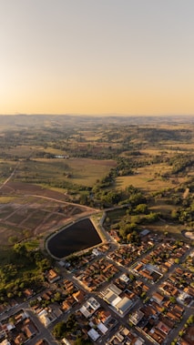 Aerial view of a landscape showcasing a sprawling suburban area with neatly arranged houses, bordered by green fields and patches of woodland. The horizon extends to distant hills under a clear, golden sky, indicating a warm, sunny day.