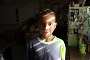 A young boy stands indoors, partially lit by sunbeam patterns across his face and shirt. Surrounding him are various personal photos pinned to a board, a variety of objects, and some visible plants, contributing to a cozy, homey environment.